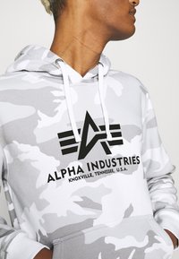 Szürke terepmintás kapucnis pulóver puha anyagból, elől zsebbel, húzózsinórral ellátott kapucnival és fekete logóval, amin az "ALPHA INDUSTRIES" felirat található.