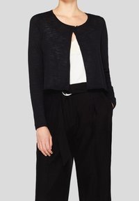 Svart kortärmad kofta, mjukt texturerad tyg, rund halsringning, öppning framtill med en enda knappknäppning, bärs över en vit topp och svarta högmidjade byxor.