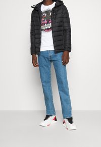 Giacca a vento nera con cappuccio, T-shirt grafica bianca, jeans azzurri chiari e sneakers bianche con dettagli rossi. Posizione in piedi, sfondo neutro.