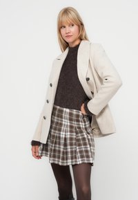 Manteau en mélange de laine beige avec des boutons noirs, associé à un pull en laine foncé et fuzzy, et une jupe plissée en tartan aux tons marron et blanc.
