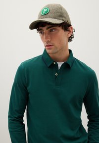 Grünes langärmeliges Polohemd mit Kragen, der mit einem dunklen Besatz und Knöpfen versehen ist. Das Modell trägt eine braune, mit Fischgrätmuster versehene Mütze mit einem Logo.