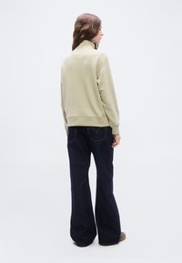 Γυναίκα με την πλάτη γυρισμένη φοράει μπεζ μακρυμάνικη μπλούζα με ψηλό λαιμό και σκούρο μπλε τζιν με φαρδιά πόδια, σε έναν απλό λευκό φόντο.