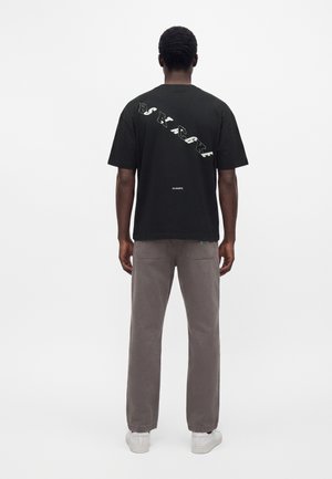 Mand set bagfra iført en sort t-shirt med diagonalt hvid tekst og grå bukser mod en ensfarvet hvid baggrund.