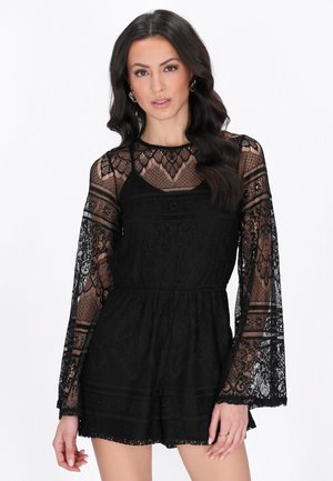 Nainen, jolla on pitkät tummat hiukset ja musta pitsinen pitkähihaisen haalari, jossa on vyötäröä korostava malli ja kukkakuvioita, seisoo valkoista taustaa vasten.