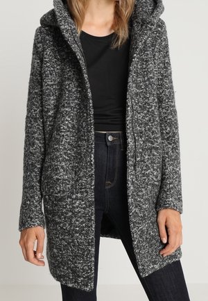 Femme porte un long cardigan à capuche gris chiné sur un top noir et un jean taille haute foncé.