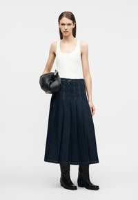 Donna che indossa un top bianco senza maniche, gonna midi in denim plissettata scura, stivali neri, che tiene una borsa nera in pelle.