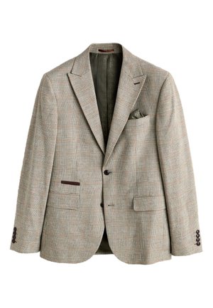 Man, van achteren gezien, draagt een beige geruit colbert en bijpassende broek tegen een neutrale beige muur.