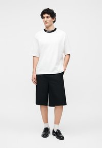 Homme debout portant un t-shirt blanc oversize avec un texte noir sur le col, un short large noir jusqu’aux genoux, des chaussettes blanches et des chaussures en cuir noires.