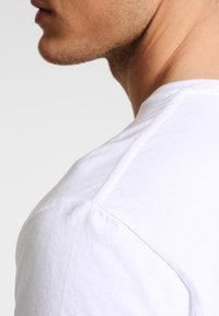 Vit bomullströja med en klassisk rundad halsringning, synliga axelsömmar och en mjuk textur, visad från en sidovinkel.