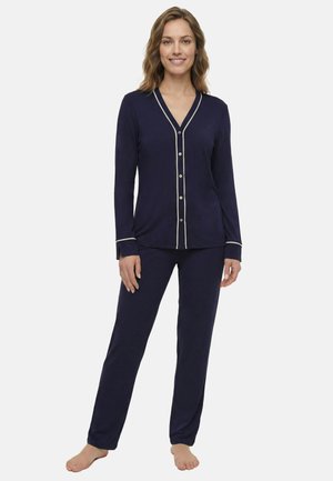 Nő áll mezítláb, tengerészkék hosszú ujjú, gombos pizsama felsőt és hozzáillő nadrágot visel, fehér szegéllyel a mandzsettán és a nyakkivágásnál.