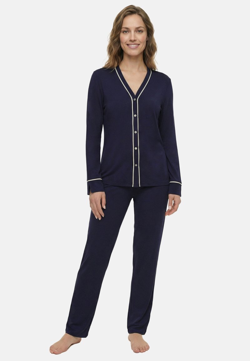 Nő áll mezítláb, tengerészkék hosszú ujjú, gombos pizsama felsőt és hozzáillő nadrágot visel, fehér szegéllyel a mandzsettán és a nyakkivágásnál.