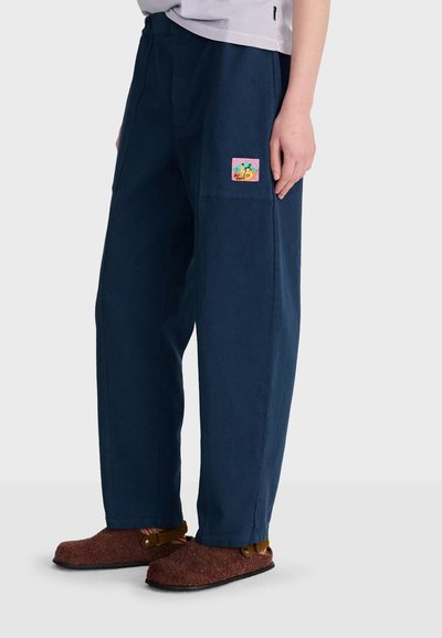Personne portant un pantalon ample bleu foncé avec un patch coloré sur la poche droite et des chaussures en feutre marron sans lacets avec des brides.