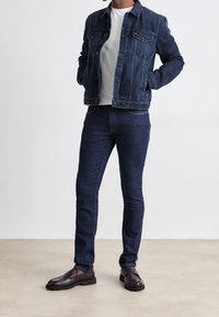 Chaqueta vaquera con bolsillos en el pecho, llevada sobre una camiseta gris, combinada con vaqueros pitillo azul oscuro y mocasines marrones con suela texturizada.