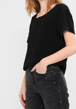 Woman wearing a black short-sleeve top and black jeans with small rhinestone embellishments, her right hand resting in her pocket.