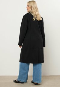 Abrigo negro largo con un diseño estructurado, que presenta hombros entallados y detalles en las costuras. Combinado con unos pantalones vaqueros de pierna ancha en un tono azul claro y calzado negro.