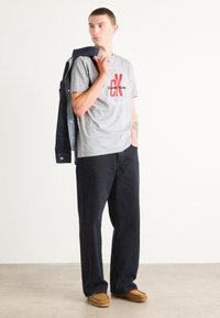 Gray cotton t-shirt featuring a red Calvin Klein logo, paired with navy loose-fit pants and a denim jacket draped over the shoulder.