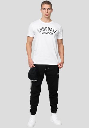 Nuori mies seisoo, yllään valkoinen "Lonsdale London" -t-paita, mustat juoksuhousut valkoisin raidoin, valkoiset tennarit ja kädessään musta lippalakki.