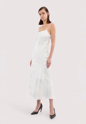 Femme portant une robe midi blanche sans manches à fines bretelles et des talons hauts noirs, debout devant un fond clair uni.