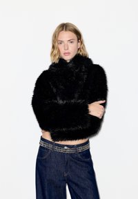 Junge Person mit kurzem blondem Haar, die einen schwarzen, flauschigen Pullover und dunkelblaue Jeans mit einem Kettengürtel trägt, steht vor einem weißen Hintergrund.