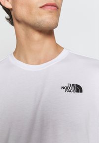 Bílý krátký rukáv tričko s kulatým výstřihem, na kterém je černé logo v oblasti levého prsa. Měkká látka, hladká textura.