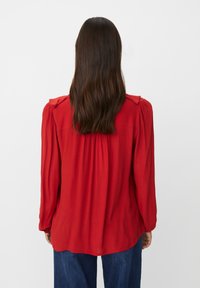 Rote Bluse mit langen Ärmeln und Schulterdetails, leicht gerafftem Rücken und weichem, strukturiertem Stoff. Kombiniert mit blauen Jeans.