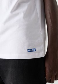 Bílý bavlněný tričko s modrým logem "HUGO" na lemu, které má klasický zaoblený límec a krátké rukávy, doplněn�é černými džíny.