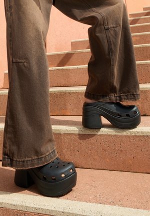 Personne portant un pantalon large marron et des sabots à plateforme noirs montant des escaliers en pierre de terre cuite.