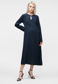 Blonda sieviete, tērpusies tumši zilā garā piedurknē midi kleitā ar atslēgas formas izgriezumu un melnām augstpapēžu sandalēm, stāvot uz vienkārša fona.