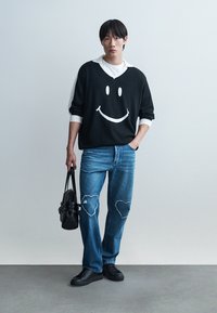 Junger Mann mit schwarzem Pullover mit weißem Smiley, weißem Hemd, blauer Jeans mit Herz-Patches, schwarzen Sneakern, der eine schwarze Handtasche hält und vor einer grauen Wand steht.