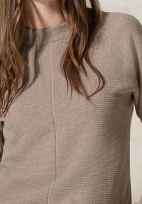 Beige texturerad tröja med hög halsringning och vertikala sömdetaljer, tillverkad av ett mjukt, stickat material, lämplig för avslappnad användning.