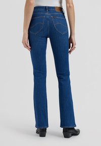 Femme portant un jean bootcut bleu et des bottes noires à talons, debout devant un fond uni, vue de dos.
