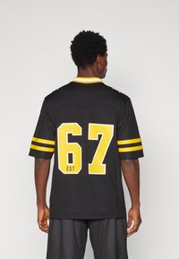 Fekete sportpóló sárga csíkokkal, a hátsó részen vastag betűkkel "67" számmal. Könnyű anyagból készült, laza szabású és kerek nyakkal.