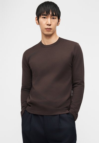 Άνδρας με κοντά μαύρα μαλλιά, που φοράει καφέ μακρυμάνικο πουκάμισο και σκούρο παντελόνι, στέκεται με τα χέρια στις τσέπες μπροστά σε απλό λευκό φόντο.