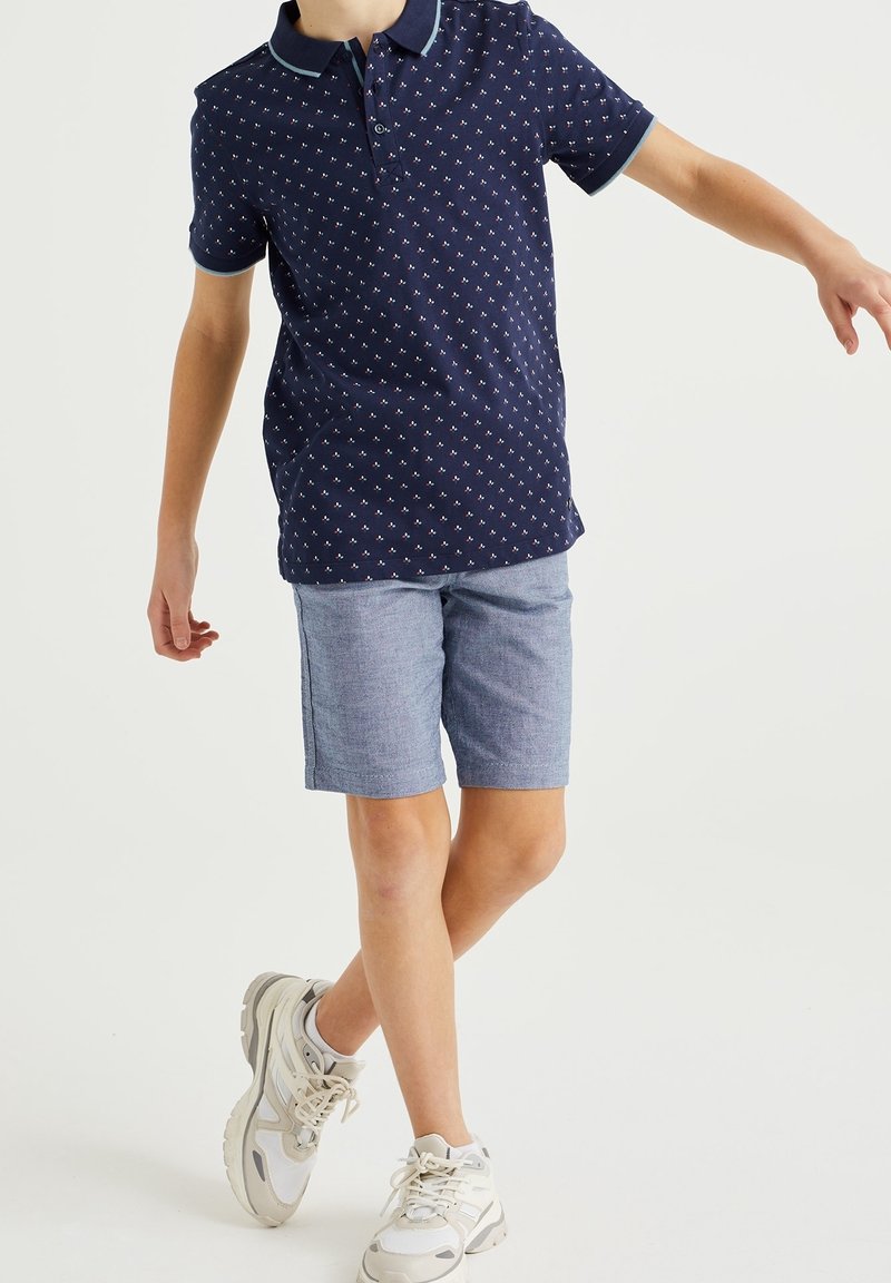 Garçon portant un polo bleu marine à motifs, un short bleu clair et des baskets blanches, debout avec une jambe croisée sur l’autre.