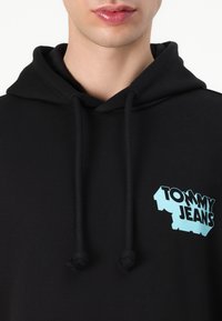 Zwarte hoodie met trekkoorden, voorzien van een lichtblauw "TOMMY JEANS" logo op de linkerkant van de borst. Zachte textuur; minimalistisch ontwerp zonder patronen.