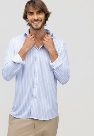 Hombre con cabello castaño claro sonríe mientras ajusta el cuello de una camisa de rayas azul claro, de pie frente a un fondo gris claro.