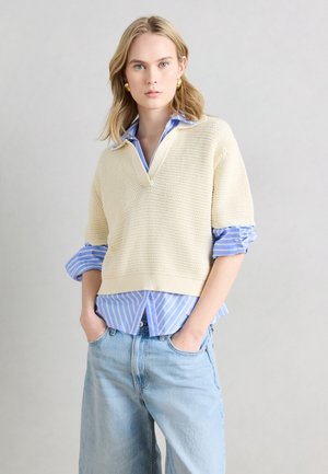 Joven mujer que lleva un suéter corto de punto color crema sobre una camisa a rayas azules con las mangas remangadas y vaqueros anchos de color azul claro, de pie con las manos en los bolsillos.