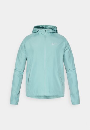 Šviesiai žalia Nike striukė su gobtuvu, užtrauktuku priekyje ir šoninėmis kišenėmis. Nėra šiurkštus paviršius ir prigludęs dizainas, ant krūtinės esantis atšvaitinis logotipas.