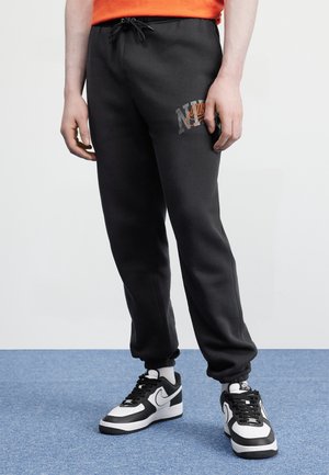 Nogi w czarnych spodniach dresowych Nike z logo na udzie, białe buty sportowe Nike z czarnymi akcentami, stojące na niebieskim dywanie.