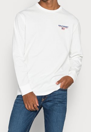 Camisa blanca de manga larga con cuello redondo, con el logo "POLO SPORT" en azul y rojo, combinada con vaqueros azules. Textura lisa.