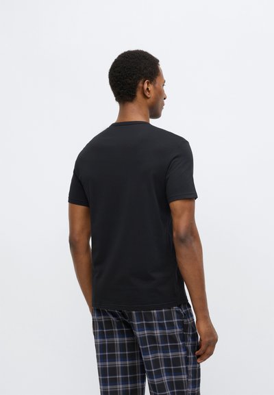 Černé tričko s krátkým rukávem z bavlny, s kulatým výstřihem. Nošeno s károvanými kalhotami, zobrazené zezadu na bílém pozadí.