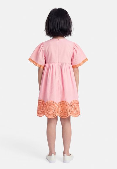 Παιδί με κοντά σκούρα μαλλιά που φοράει ένα ροζ φόρεμα με πορτοκαλί δαντέλα και λευκά παπούτσια, στέκεται γυρισμένο προς τα πίσω σε λευκό φόντο.