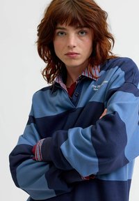 Zila un tumši zila svītrainā polo krekls ar garām piedurknēm, izgatavots no mīksta auduma ar rūtainu apkakli. Ierakstīta logo izšuvums uz krūtīm.