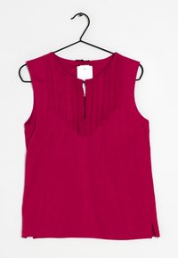 Haut sans manches fuchsia avec un col plissé, présentant une fente à l'avant et une étiquette attachée, fabriqué en tissu doux. Suspendu à un crochet noir.