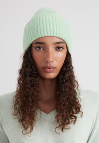 Bonnet tricoté en vert menthe clair, avec une texture côtelée et un sommet pointu. Le tissu est doux et extensible, conçu pour la chaleur.