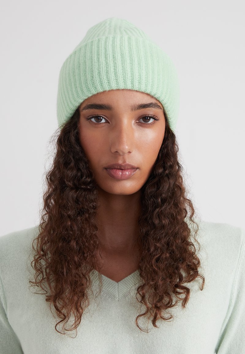 Bonnet tricoté en vert menthe clair, avec une texture côtelée et un sommet pointu. Le tissu est doux et extensible, conçu pour la chaleur.