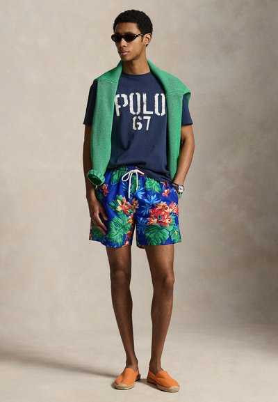 Άνδρας που φοράει γυαλιά ηλίου, μπλε μπλούζα "POLO 67", πράσινο πουλόβερ περασμένο στους ώμους, μπλε φλοράλ σορτς, πορτοκαλί slip-on παπούτσια, στέκεται με τα χέρια στις τσέπες.
