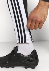 Svart fotbollssko med texturerad yta, tillsammans med vit strumpa och svarta byxor med tre vertikala vita ränder längs sidorna.