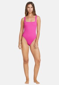 Maillot de bain une pièce rose, côtelé, avec décolleté carré et larges bretelles, présentant des découpes hautes sur les jambes et une silhouette ajustée.