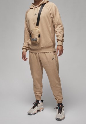 Homme portant un sweat à capuche Jordan beige et un pantalon de survêtement, avec un sac à bandoulière Jordan beige, ainsi que des chaussures de sport beige, noires et blanches, debout sur un sol gris.